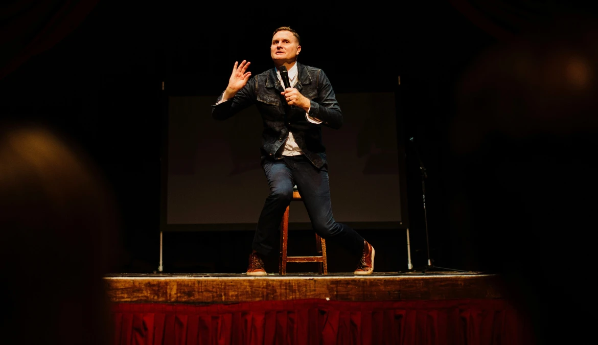 A comedian perched on a wooden stool mid-set, gesturing with a microphone in front of a projection screen