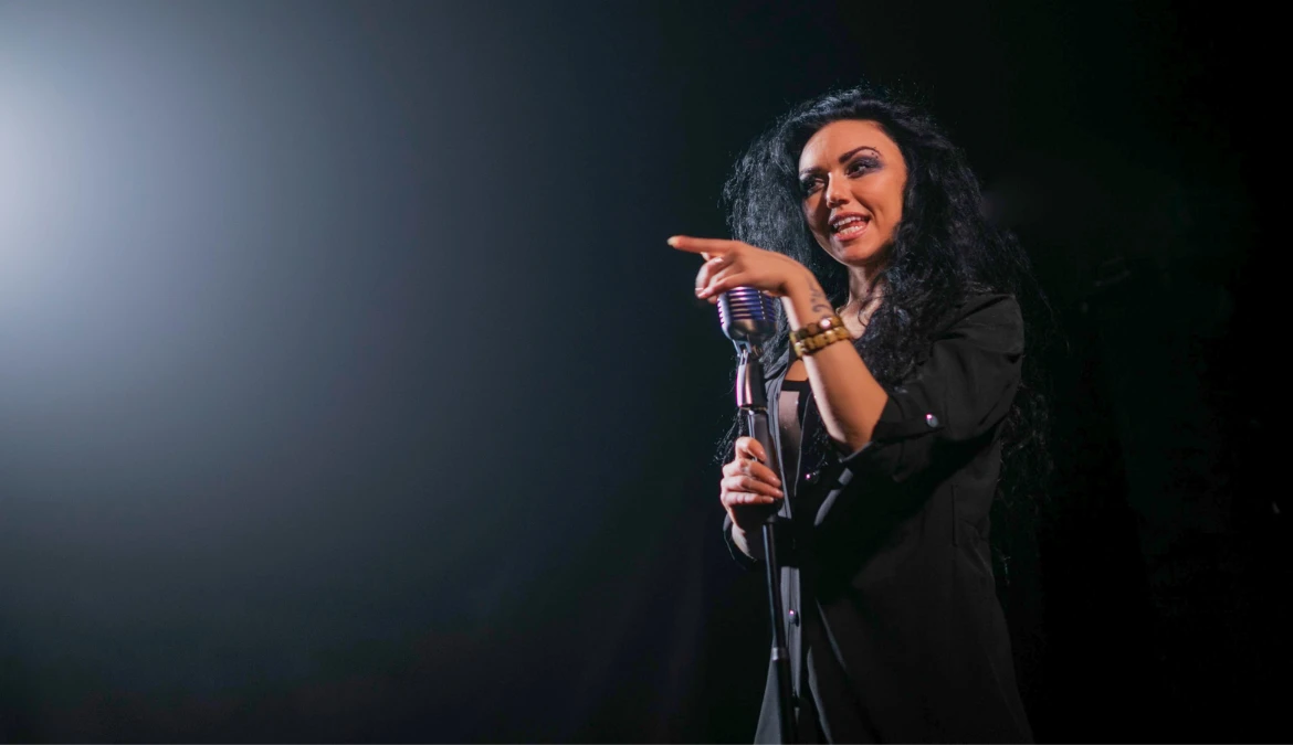 A comedian under a spotlight holding a microphone and pointing out towards the audience mid-punchline