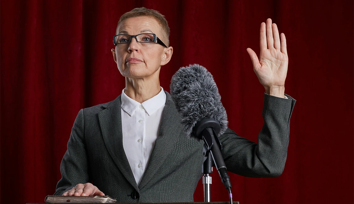 A speaker in a grey suit at a lectern, one hand raised mid-address, against a red curtain