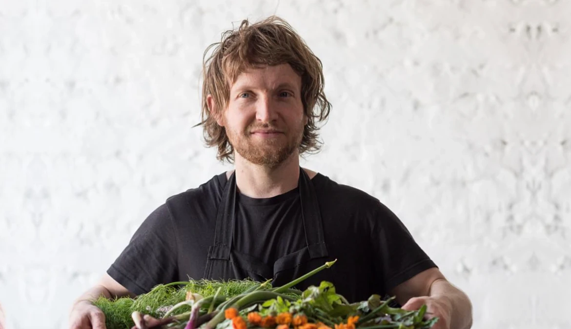 A chef in a black apron holding a crate of freshly picked herbs, spring onions and edible flowers against a whitewashed wall