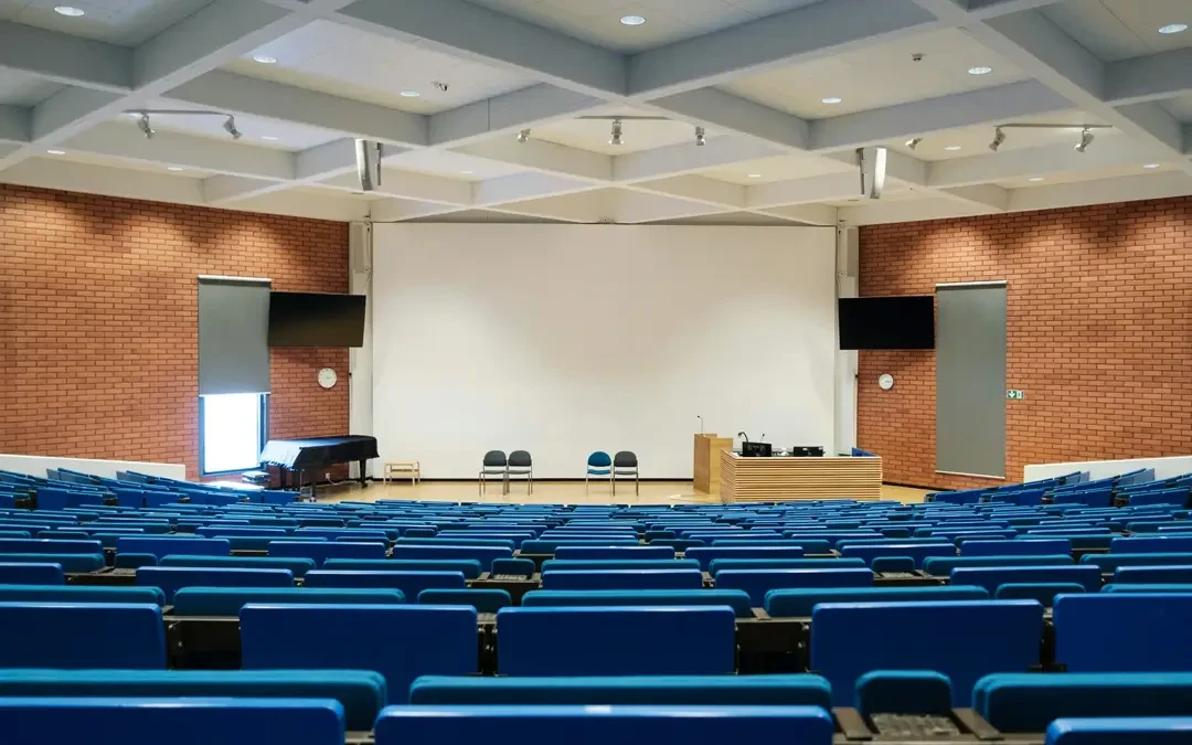 Empty university lecture hall with blue seating, a large white presentation screen, and a podium at the front.