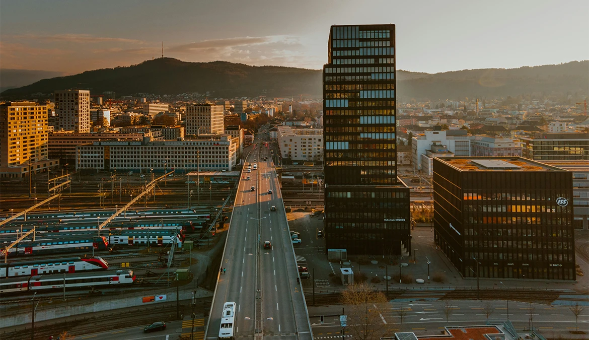 Aerial view of Zurich at golden hour, a road and railway sidings running toward the city with wooded hills behind